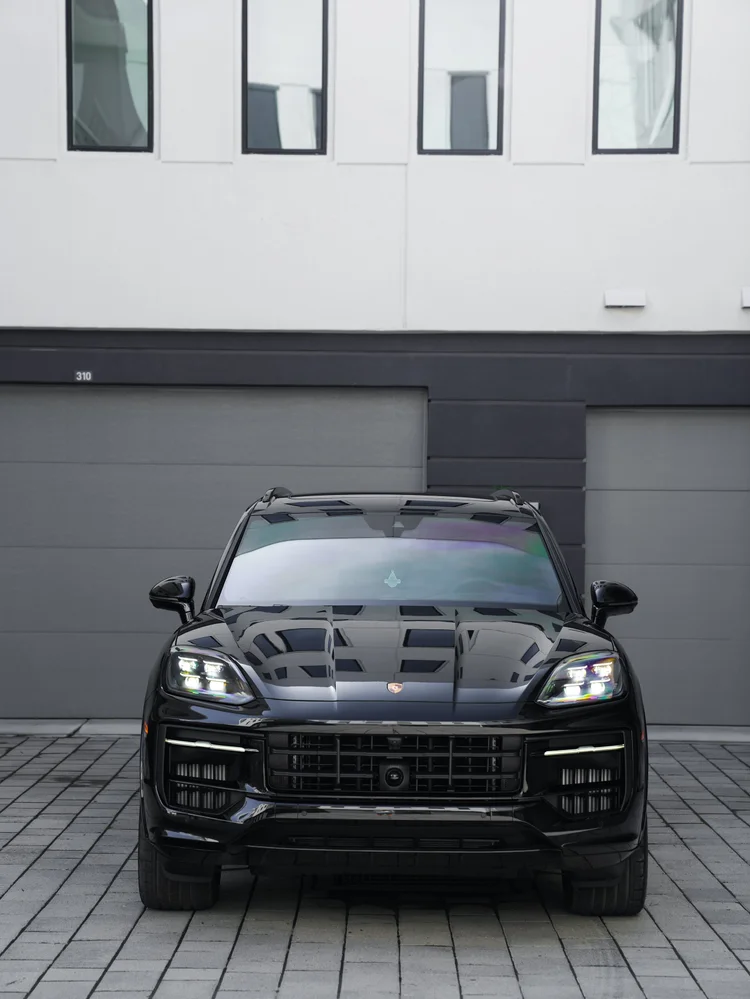 Lower straight-front view of the black Porsche Cayenne GTS with its grille, illuminated headlamps and front tyres against a light building.