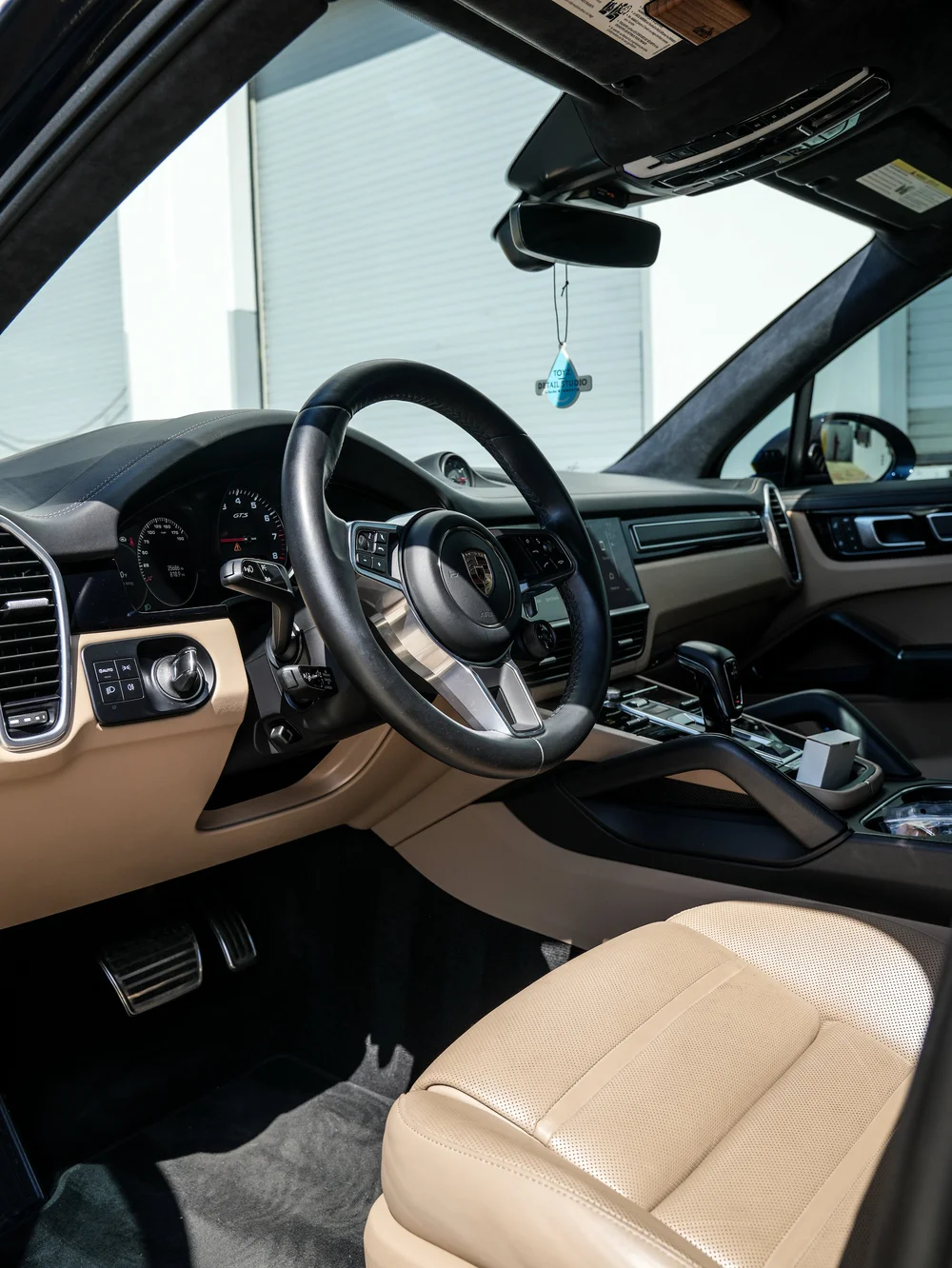 Porsche Cayenne GTS cockpit viewed through the open driver door, showing a Porsche steering wheel, tan lower trim, dark dashboard and center console.