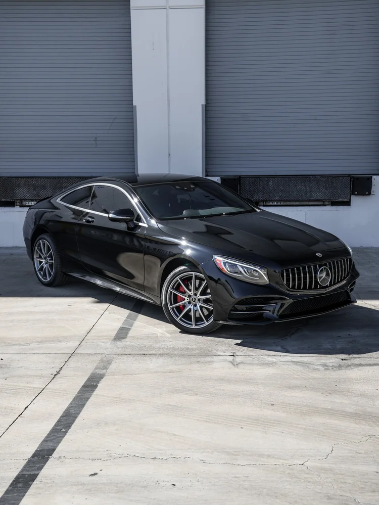 Second source photograph of the same front three-quarter composition of the black Mercedes-Benz S 560 coupe, with silver wheels, red brake calipers and bright trim beside gray roller doors.