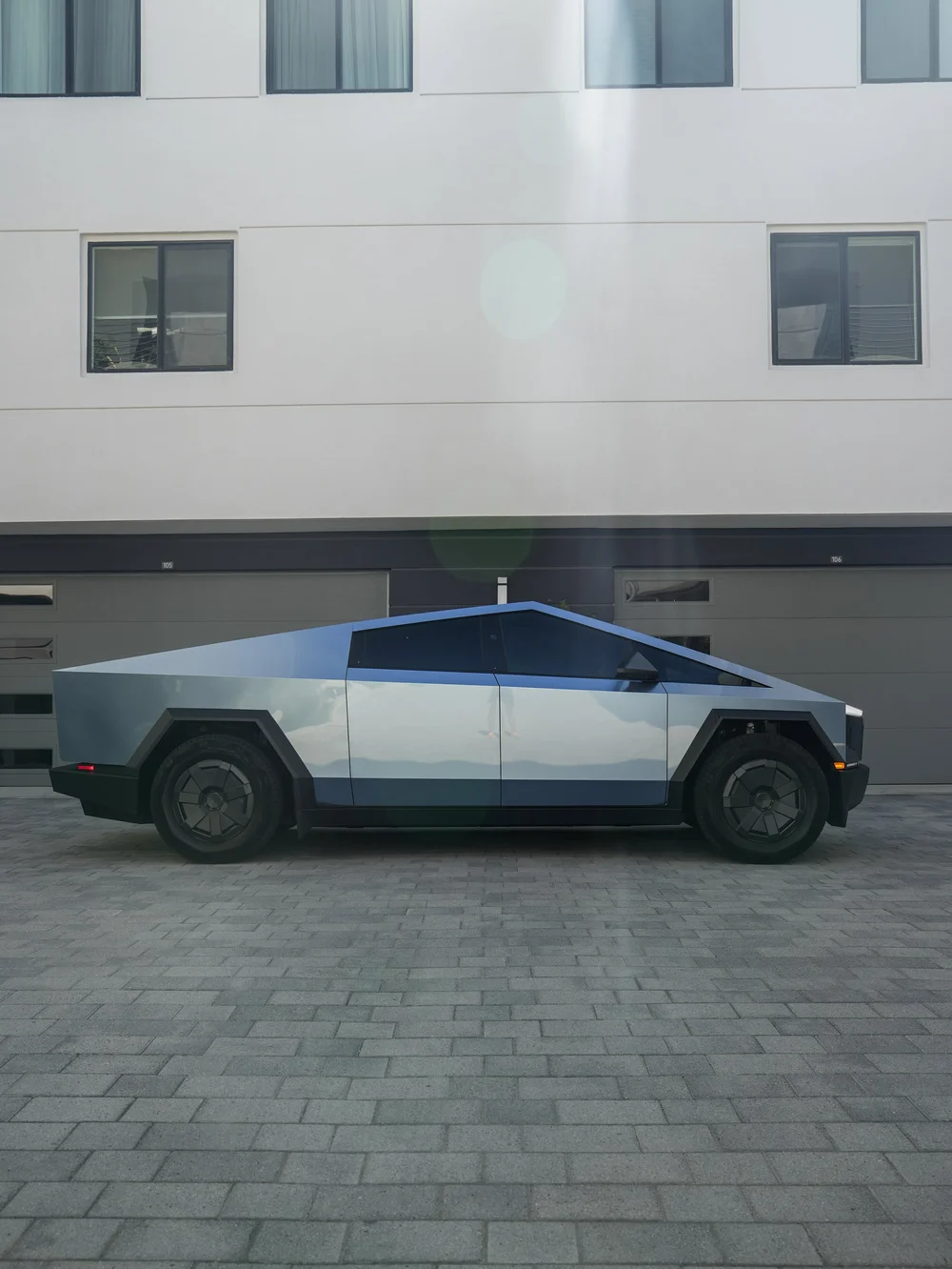 Complete side profile of the blue-gray Cybertruck facing right, with reflected clouds across the doors, dark side glass and two angular wheel covers.