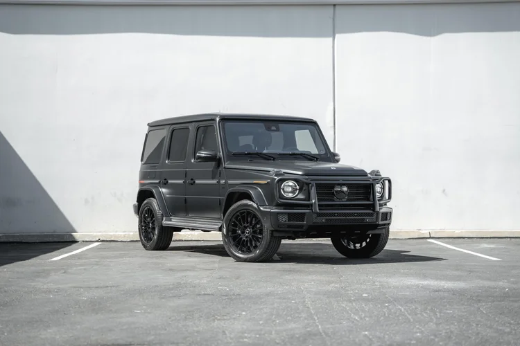 Complete front-three-quarter view of a dark grey Mercedes-Benz G 550 with black wheels and circular headlamps beside a white wall.