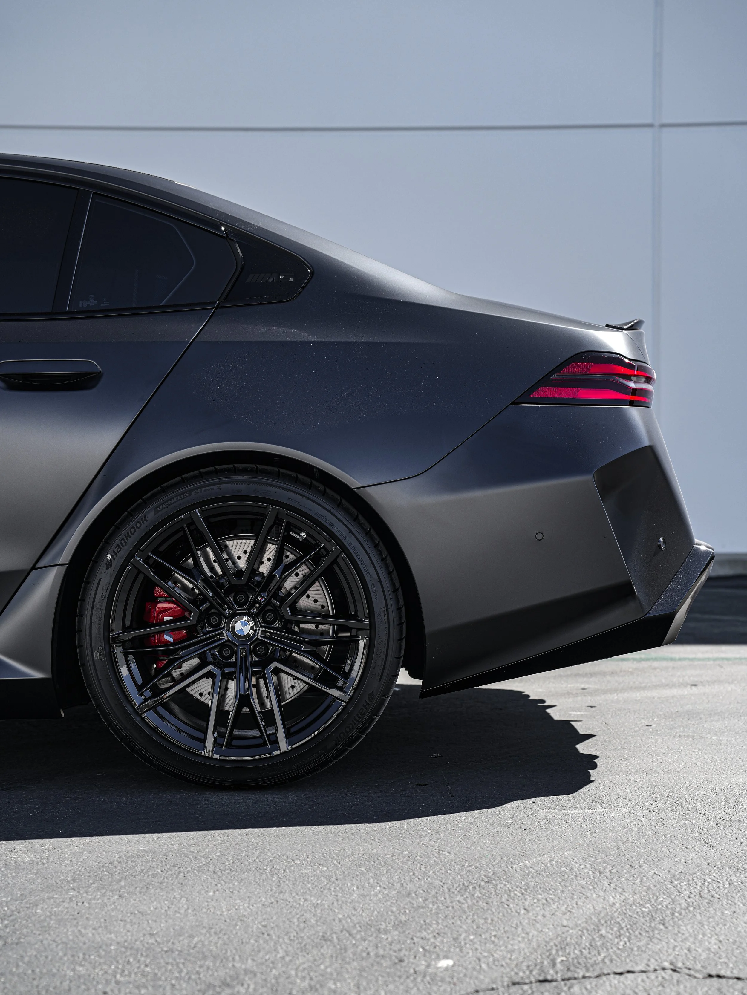 Close-up of the rear side of a black BMW sports car parked on a paved surface, showing the wheel, brake caliper, and part of the trunk with a modern building in the background.
