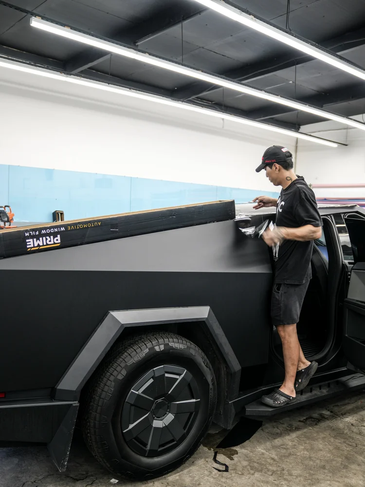 A technician working near the upper side-panel edge of the matte black Tesla Cybertruck in the studio, with an open doorway and a film box nearby.
