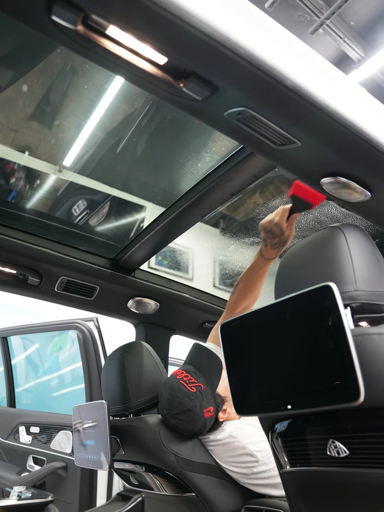 Technician working a red squeegee across wet overhead roof glass from inside the cabin beside seatback displays.