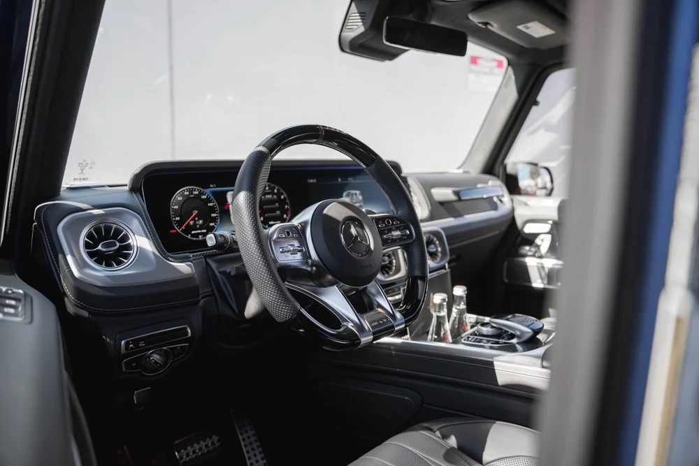 Dark G 63 cabin with circular vents, a Mercedes-star steering wheel, dashboard displays and bottles beside console controls