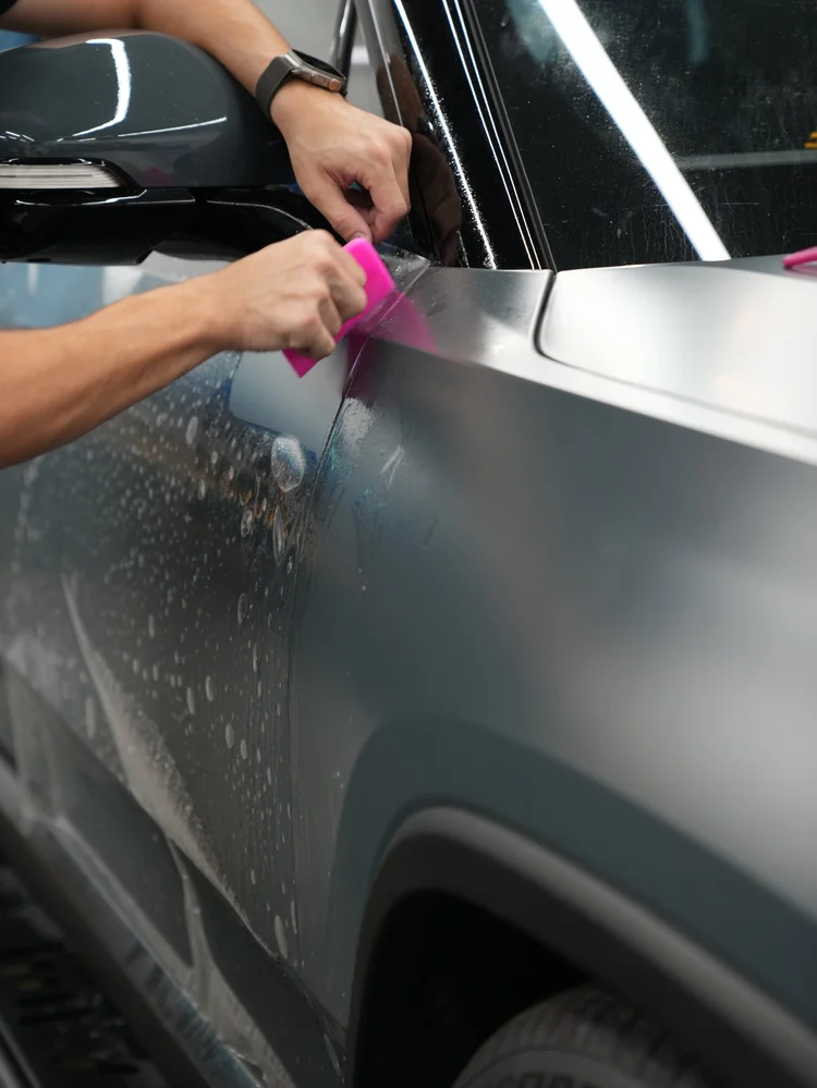Hands holding a pink squeegee and film at the Rivian R1T front-fender edge beside the side mirror, dark trim and wet glass.