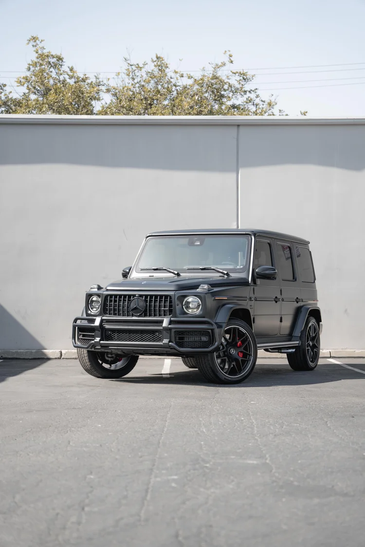 Tall complete front-three-quarter portrait of the grey G 63, with its dark wheels and circular lamps below treetops