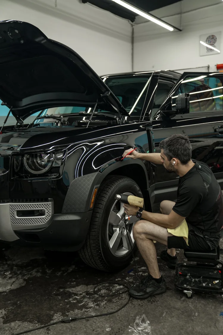 Installer using a red tool at the Defender's reflective front fender while holding a handheld nozzle tool, with the hood raised and front door open