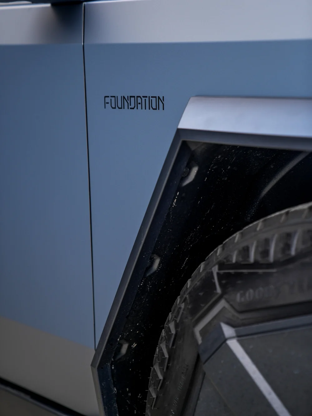 Close Tesla Cybertruck panel detail showing a closed panel seam, grey blue finish, visible FOUNDATION lettering, black wheel-arch trim and part of a tire.