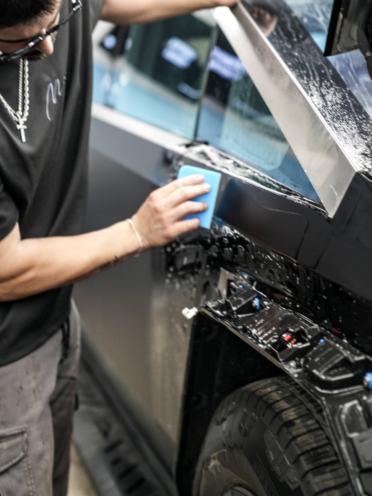 A technician using a blue squeegee on wet black film below the Tesla Cybertruck triangular side glass, with exposed mirror-mount hardware and an open panel visible.