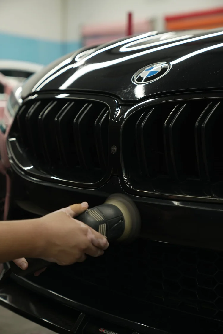 Close view of a machine polisher held against the BMW M8 front bumper below the grille and hood badge.