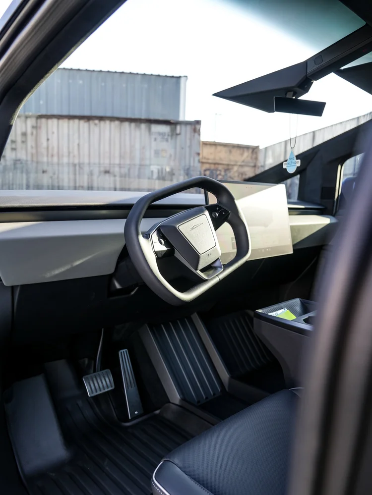 Tesla Cybertruck cockpit viewed through an open doorway, showing the steering wheel, central touchscreen, pedals and dashboard.
