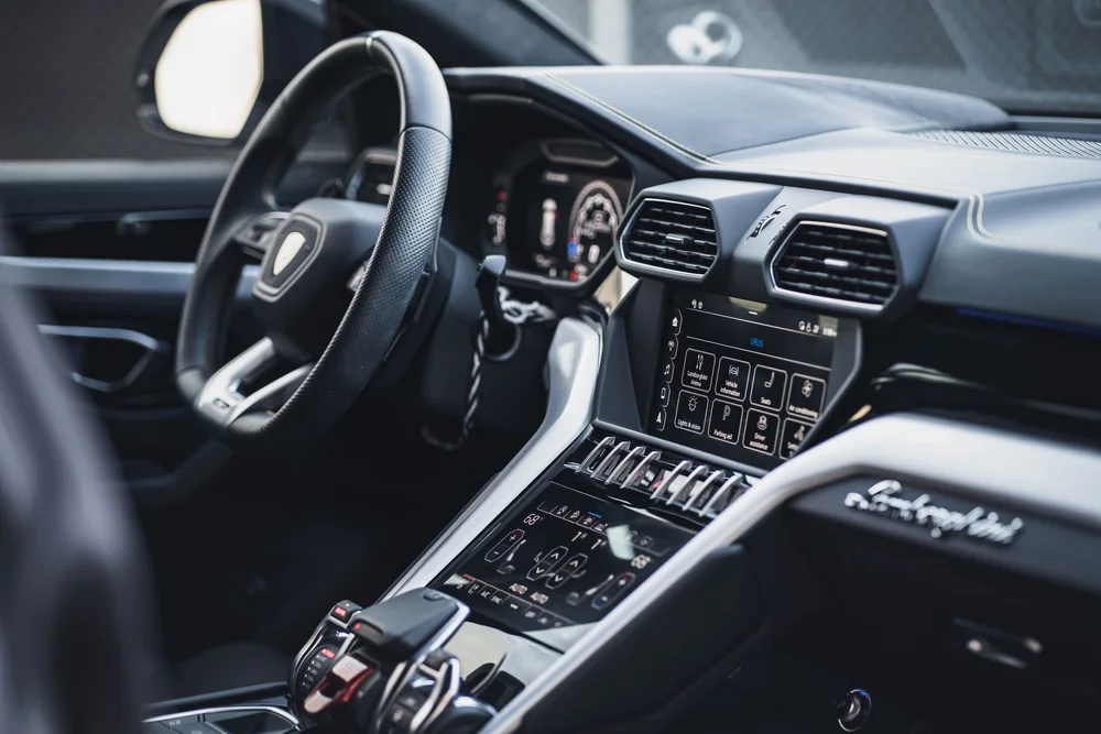 Wide dark-cabin view with the Lamborghini steering wheel, two center screens, silver-colored trim and passenger script.