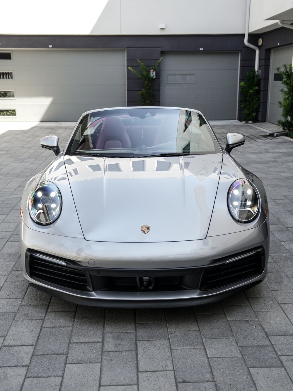 Straight front view of the silver open-roof Porsche 911 with a Porsche hood crest, oval headlights, black lower grilles and red seats visible through the windshield.