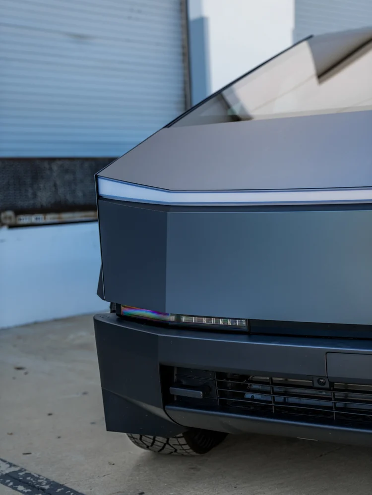 Tesla Cybertruck front detail showing the grey blue hood, light bar, headlamp, black bumper and grille.