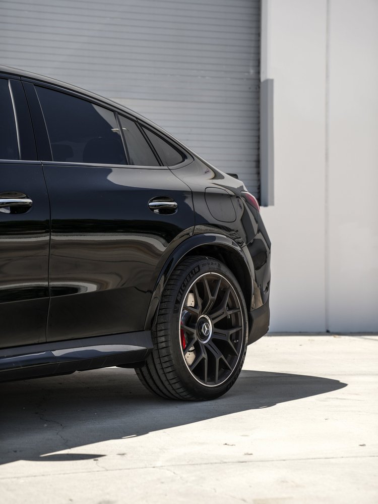 Black Mercedes-AMG GLC 63 S rear-side detail showing dark rear door glass, the sloping window line, rear wheel, red brake caliper and fuel door.