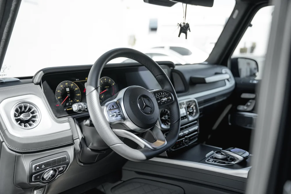 Grey-and-black G 550 cabin with a Mercedes-star steering wheel, dashboard screens, circular vents and center-console controls