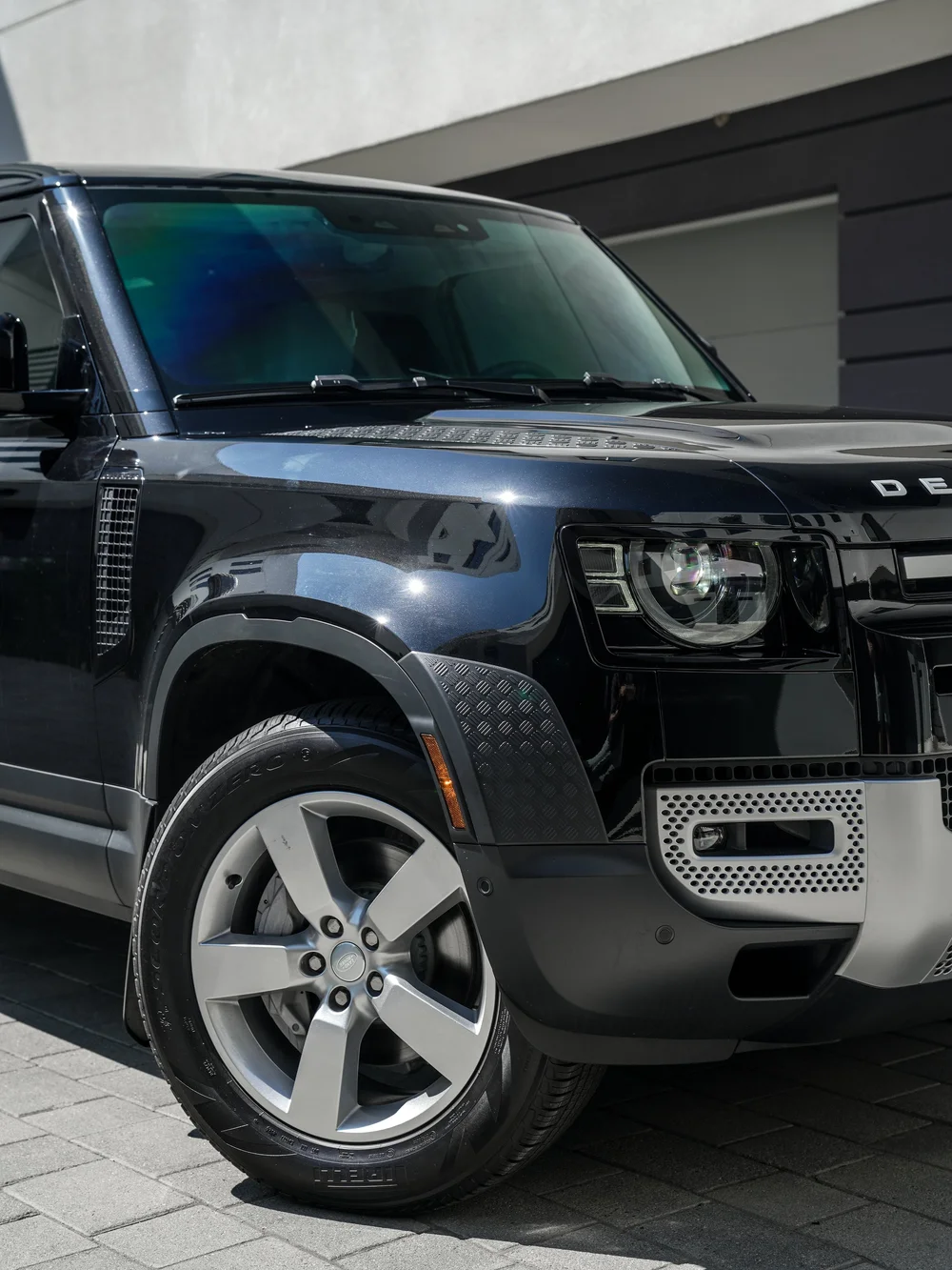 Close front corner with a silver five-spoke wheel, textured wheel-arch trim and a circular headlamp beside the cropped Defender grille