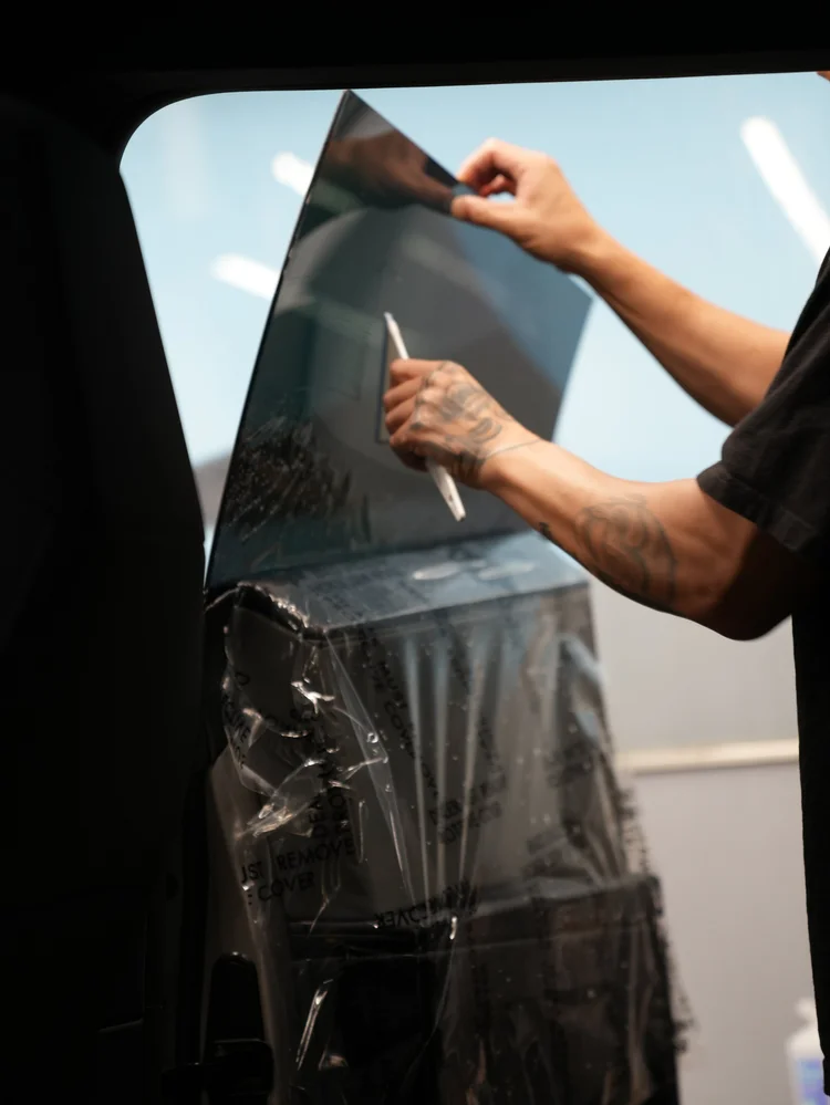 A hand pressing a white-handled squeegee across wet film on Tesla Cybertruck side glass, with an open door and protective plastic below.