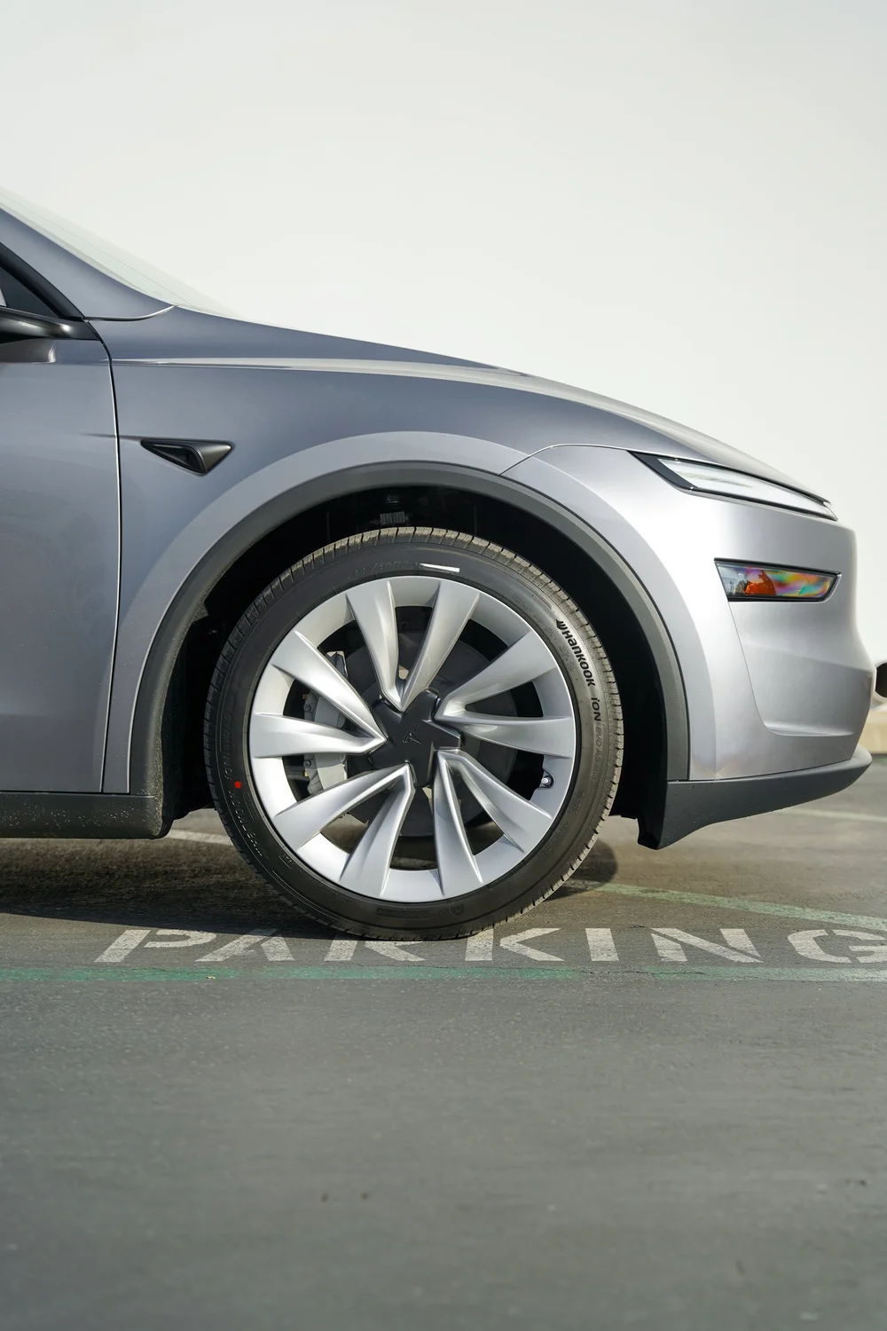Close-up of the Tesla Model Y front wheel and tyre with the gray fender, headlamps, side camera trim and lower door edge.