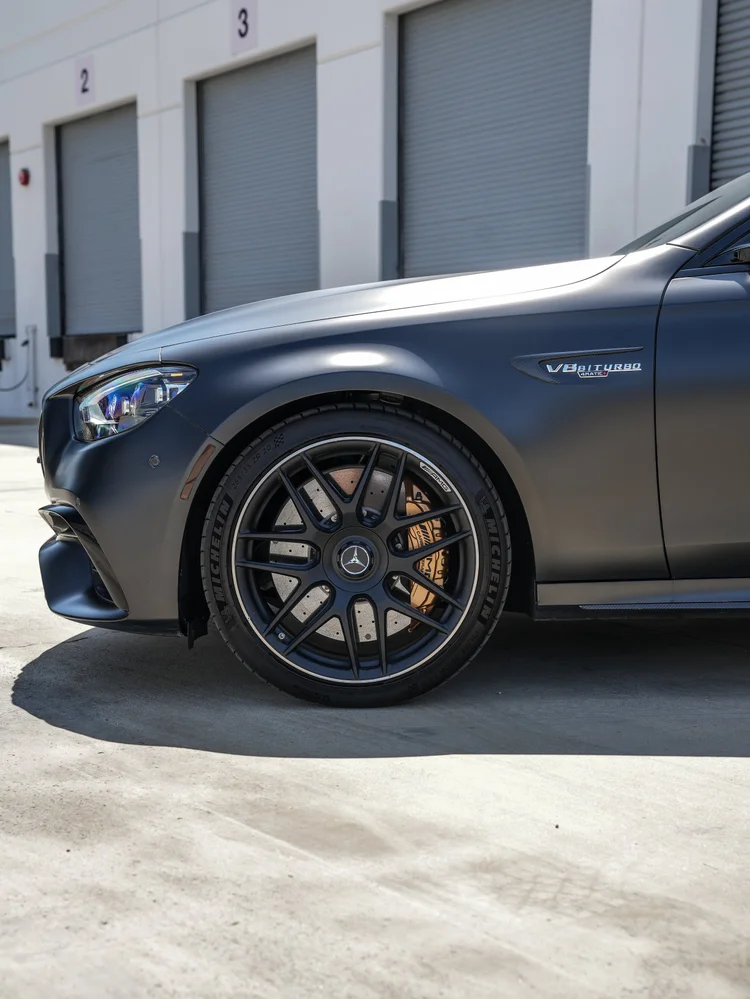 Mercedes-AMG E 63 S front-wheel and fender detail with a headlamp, yellow-gold caliper, matte-looking hood edge and visible V8 BITURBO 4MATIC+ side badge.