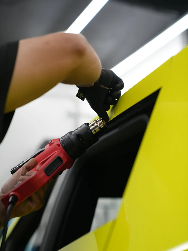 A gloved hand and red heat tool working beside yellow vinyl at a narrow black opening in an upright Tesla Cybertruck panel.