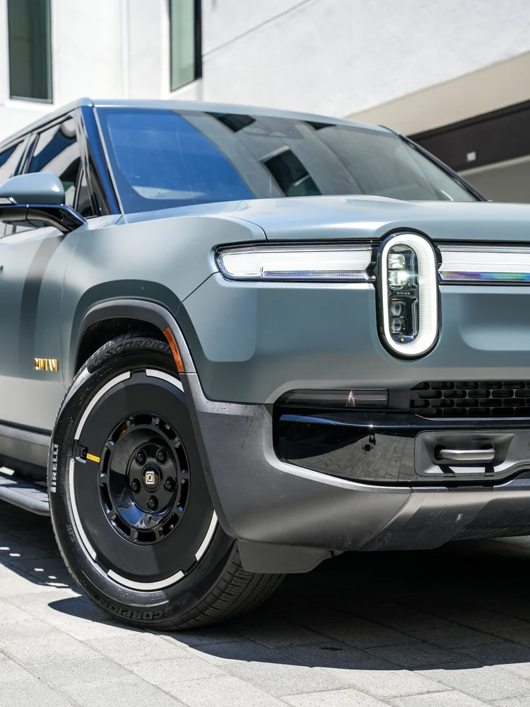 Rivian R1T front-corner detail showing an illuminated oval lamp, horizontal light bar, satin body panel, front wheel and black bumper.
