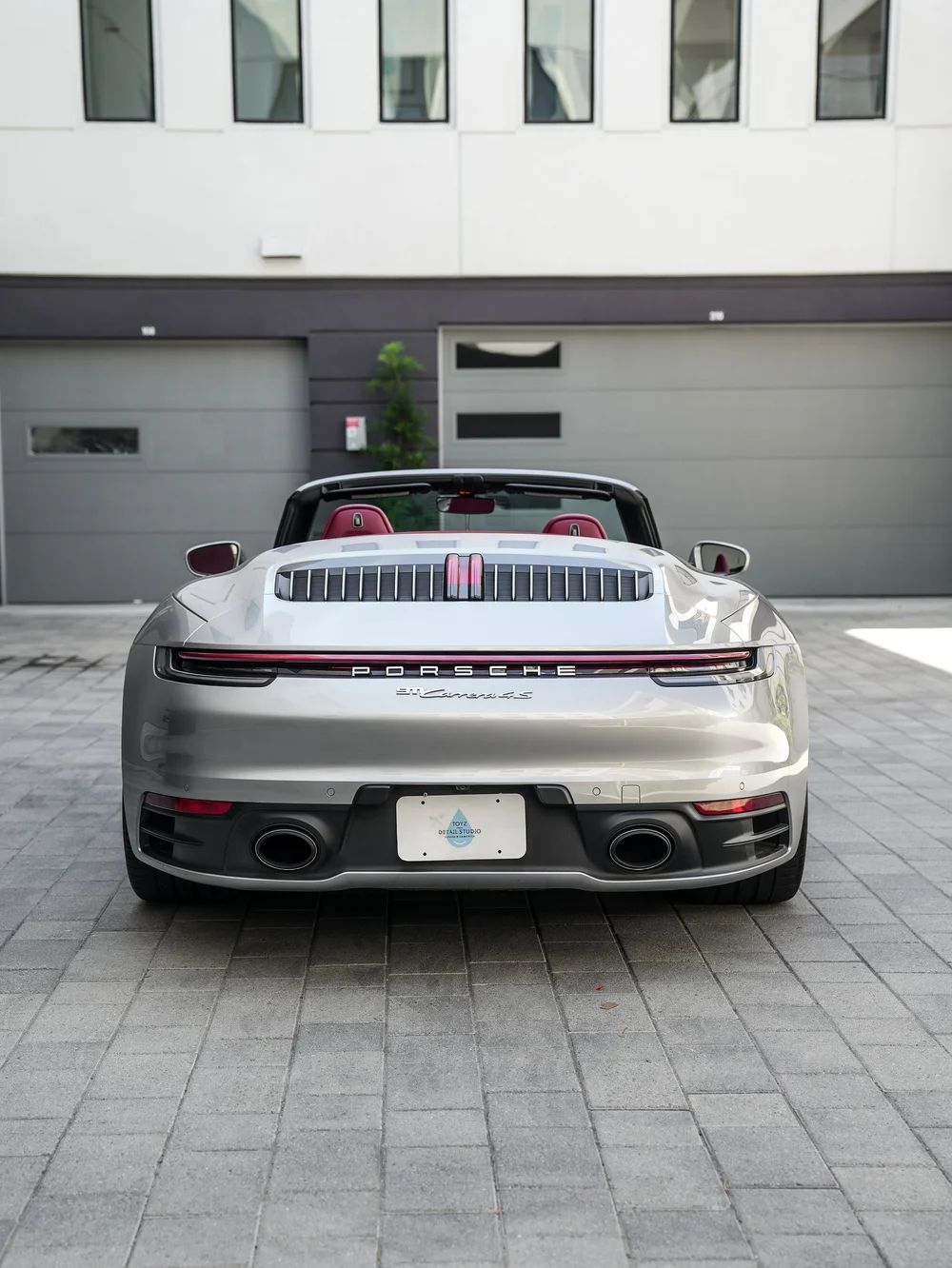 Straight rear view of the silver open-roof Porsche 911 showing red seats, vertical grille slats, a red lamp strip, Porsche lettering, a visible 911 Carrera 4 S badge and twin round exhaust outlets.