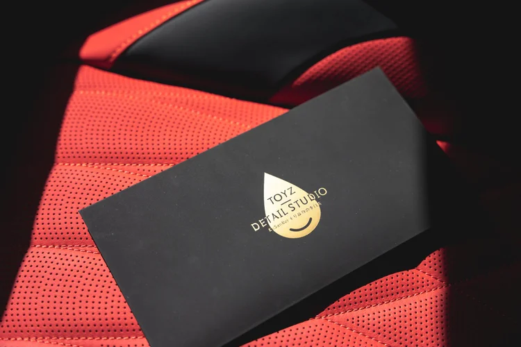 Black Toyz Detail Studio logo folder resting on a perforated red seat beside a black seat inset