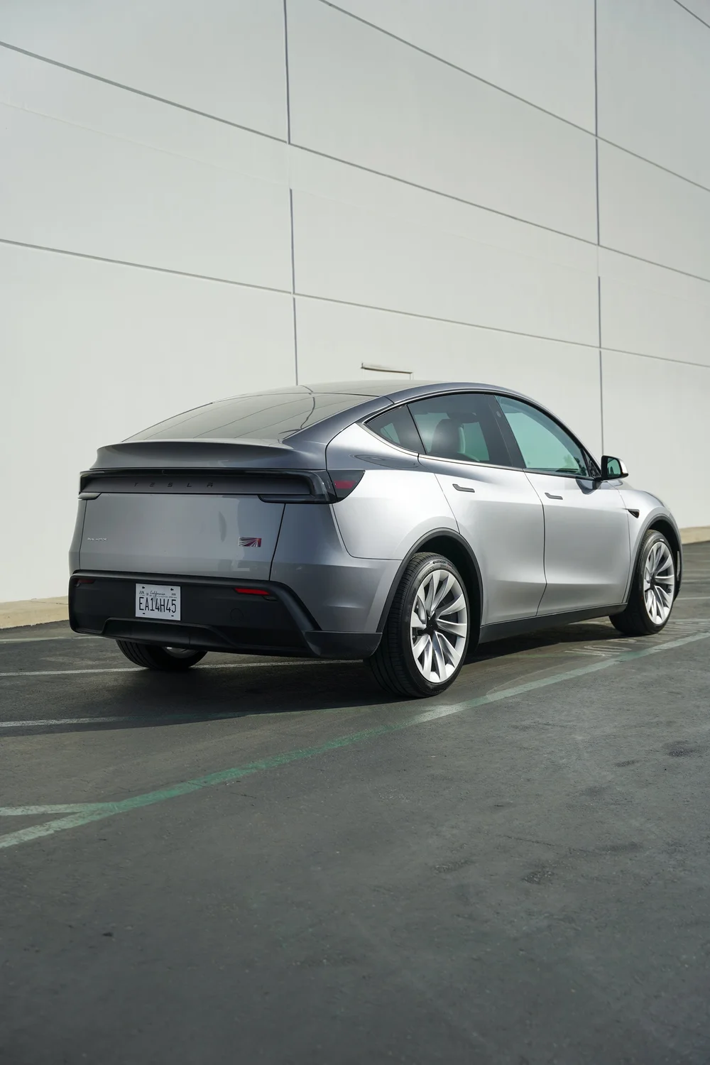 Rear three-quarter view of the gray Tesla Model Y beside a white wall, showing the liftgate, side glass, doors and silver wheels.