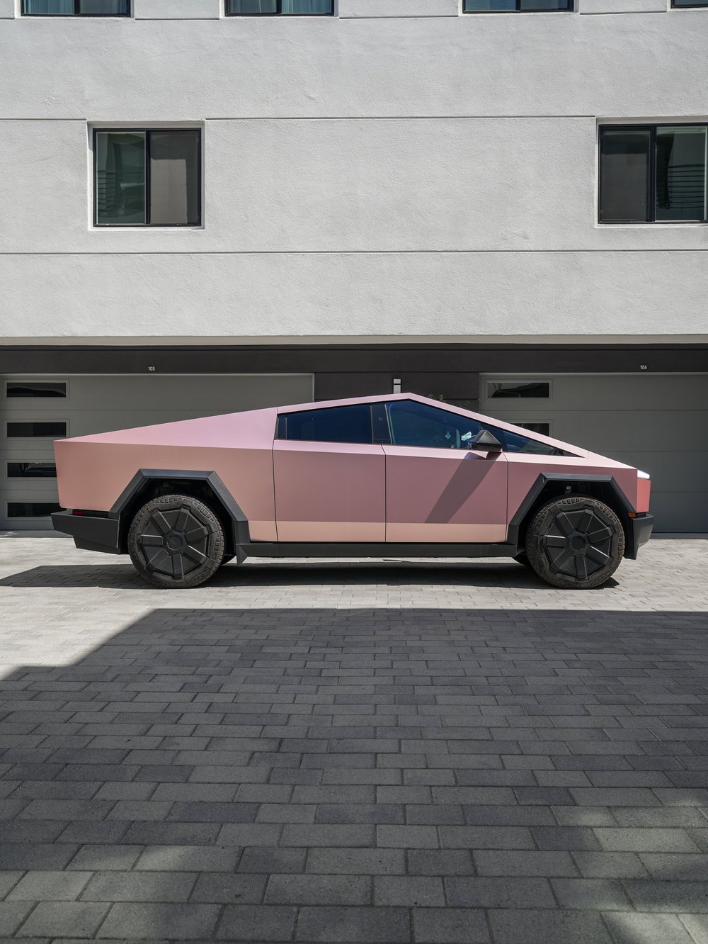 Complete side profile of the pink-gold Cybertruck with its triangular roofline, dark side windows, black wheel arches and angular wheels.