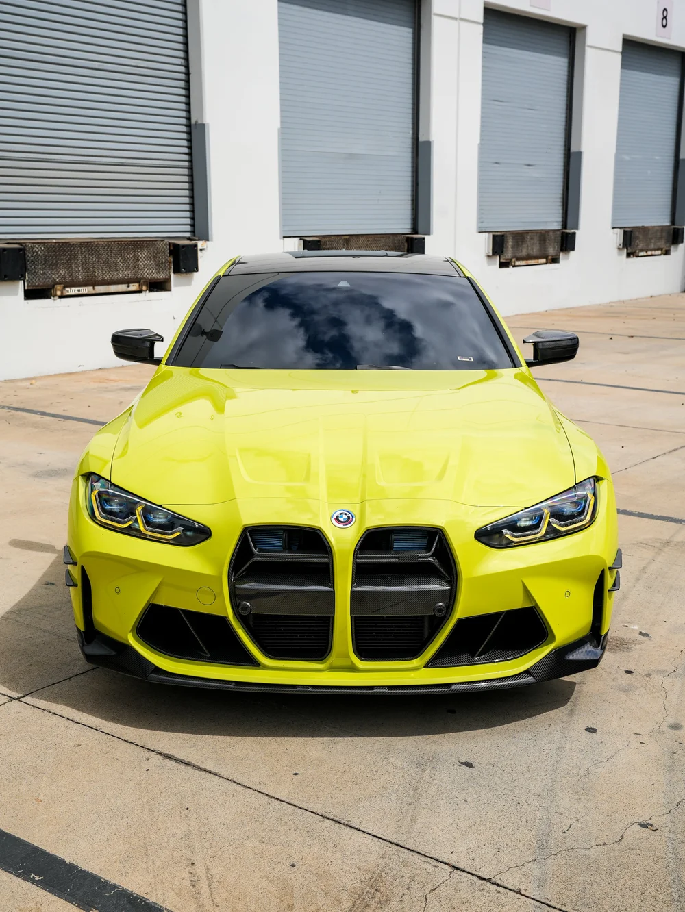 Straight front view of the yellow-green BMW M4 showing symmetrical headlights, tall black kidney grilles, hood reflections, the dark roof and both mirrors.