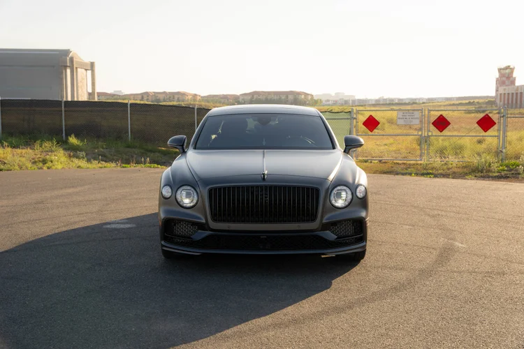 Centered front view of the sedan, its mirrors and long shadow on the paved lot.