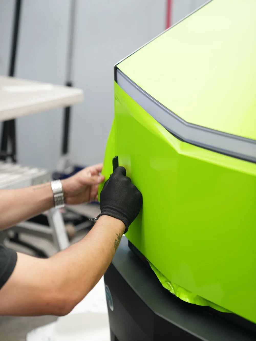Gloved hand working a small black squeegee over green vinyl on the Cybertruck front panel below the lamp strip, with loose film along the lower edge.