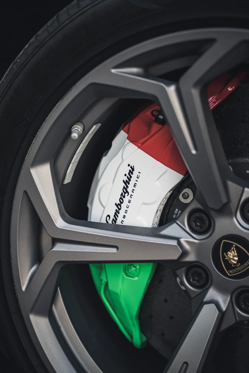 Green, white and red caliper with visible Lamborghini lettering behind a gray geometric wheel and gold shield center cap.
