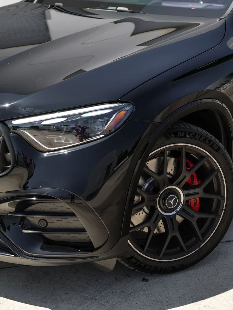 Close Mercedes-AMG GLC 63 S front-corner view showing a headlight, reflective black hood, dark multi-spoke wheel, drilled brake disc and red brake caliper.