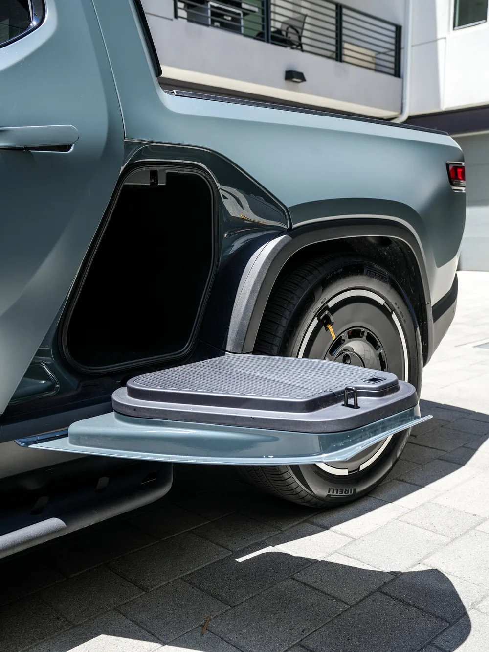Rivian R1T open side-storage compartment with its lowered door beside satin bed-side panels, rear wheel and side step.