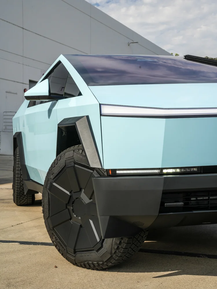 Close front three-quarter detail of the pale blue Cybertruck showing the lamp strip, dark front wheel cover, tire tread and black wheel-arch surround.