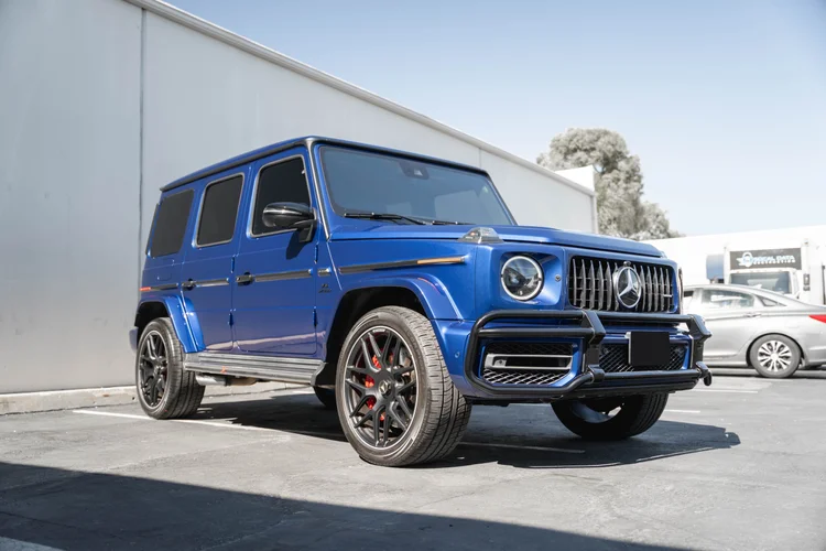 Low complete front-three-quarter view of the blue G 63 with dark wheels and red calipers, with a silver car in the background