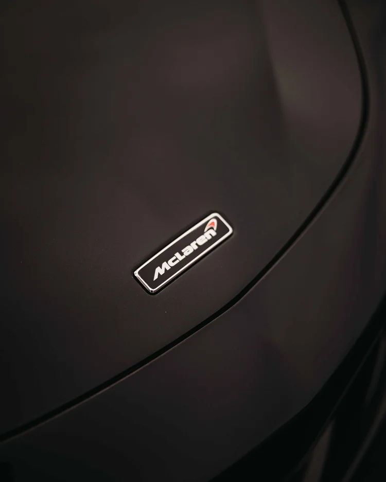 Small McLaren badge set on the curved dark hood beside a panel seam.