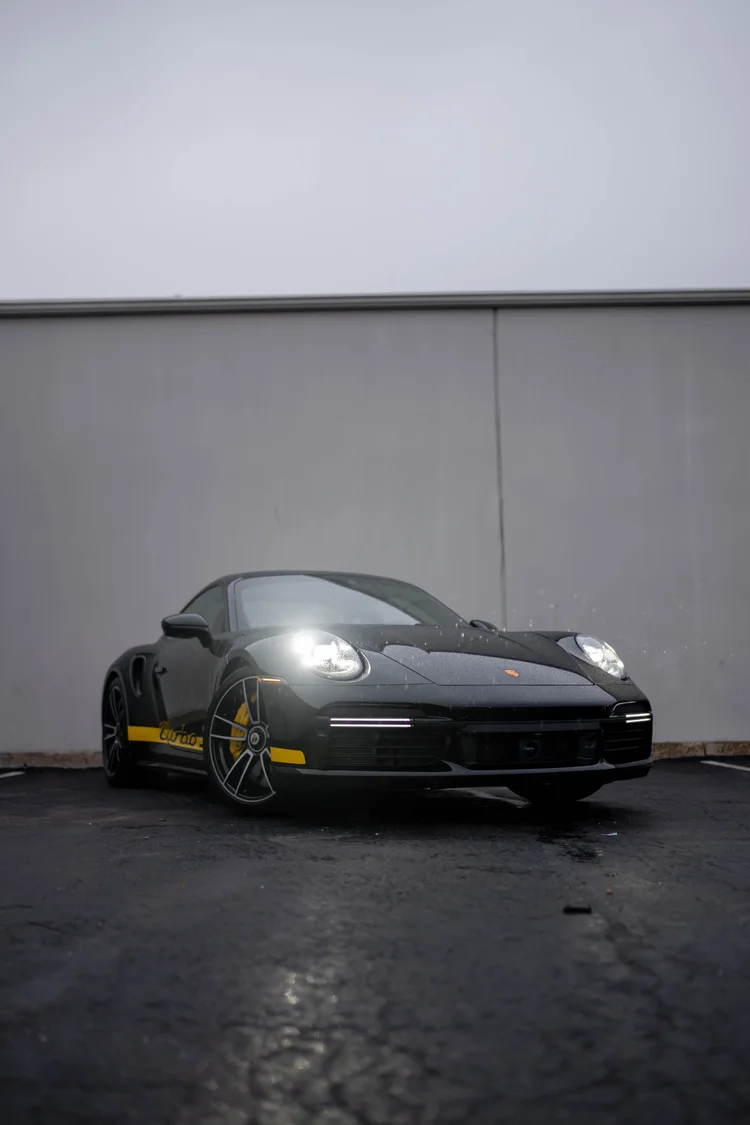 Complete front three-quarter view of a black Porsche 911 Turbo S with illuminated headlamps, yellow side graphics and yellow calipers against a gray wall.