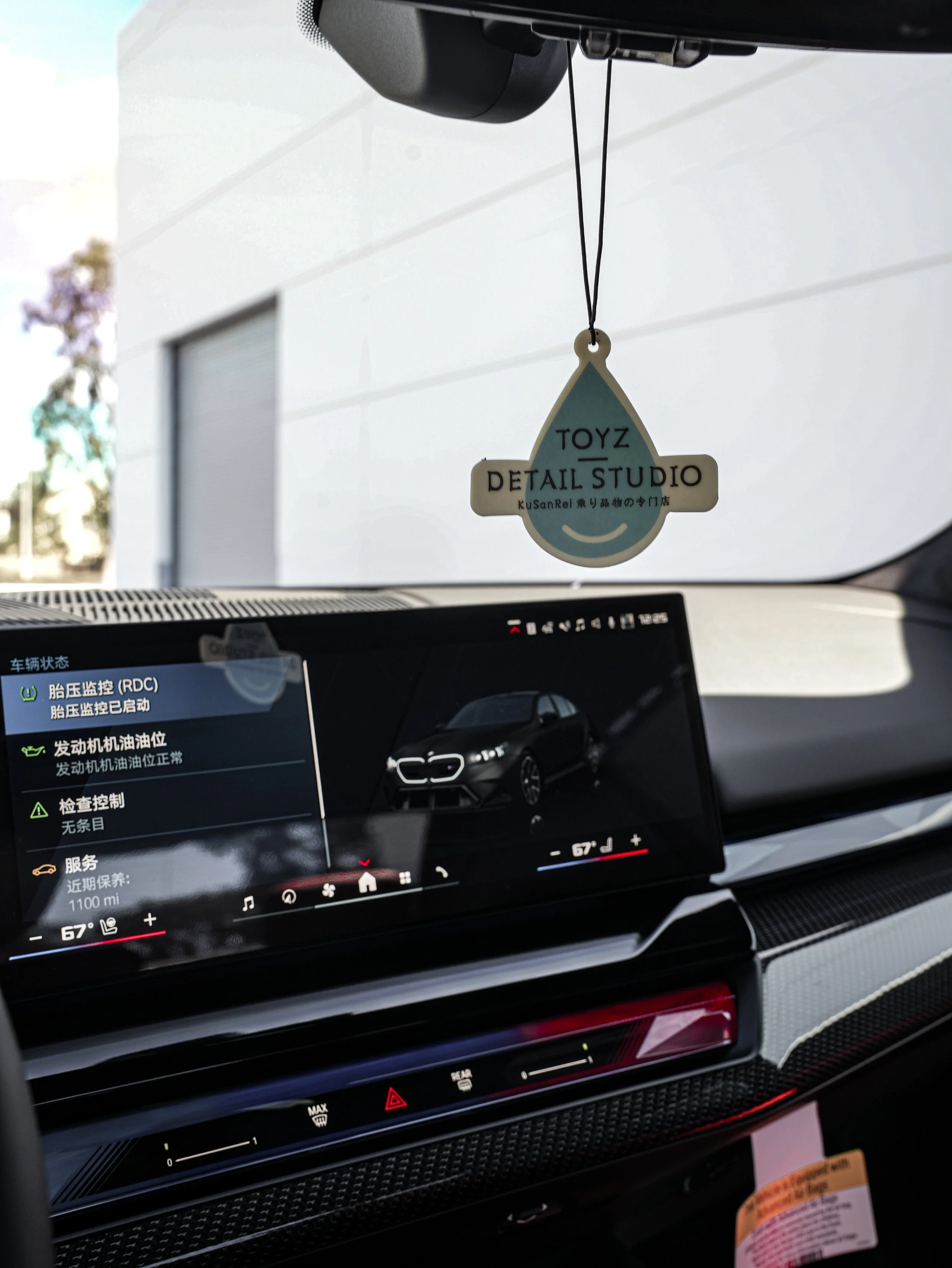 Interior of a car showing a digital dashboard with a car model display and Chinese language menu, and a hanging air freshener shaped like a teardrop with the text 'TOYZ DETAIL STUDIO KusanRei' inside.