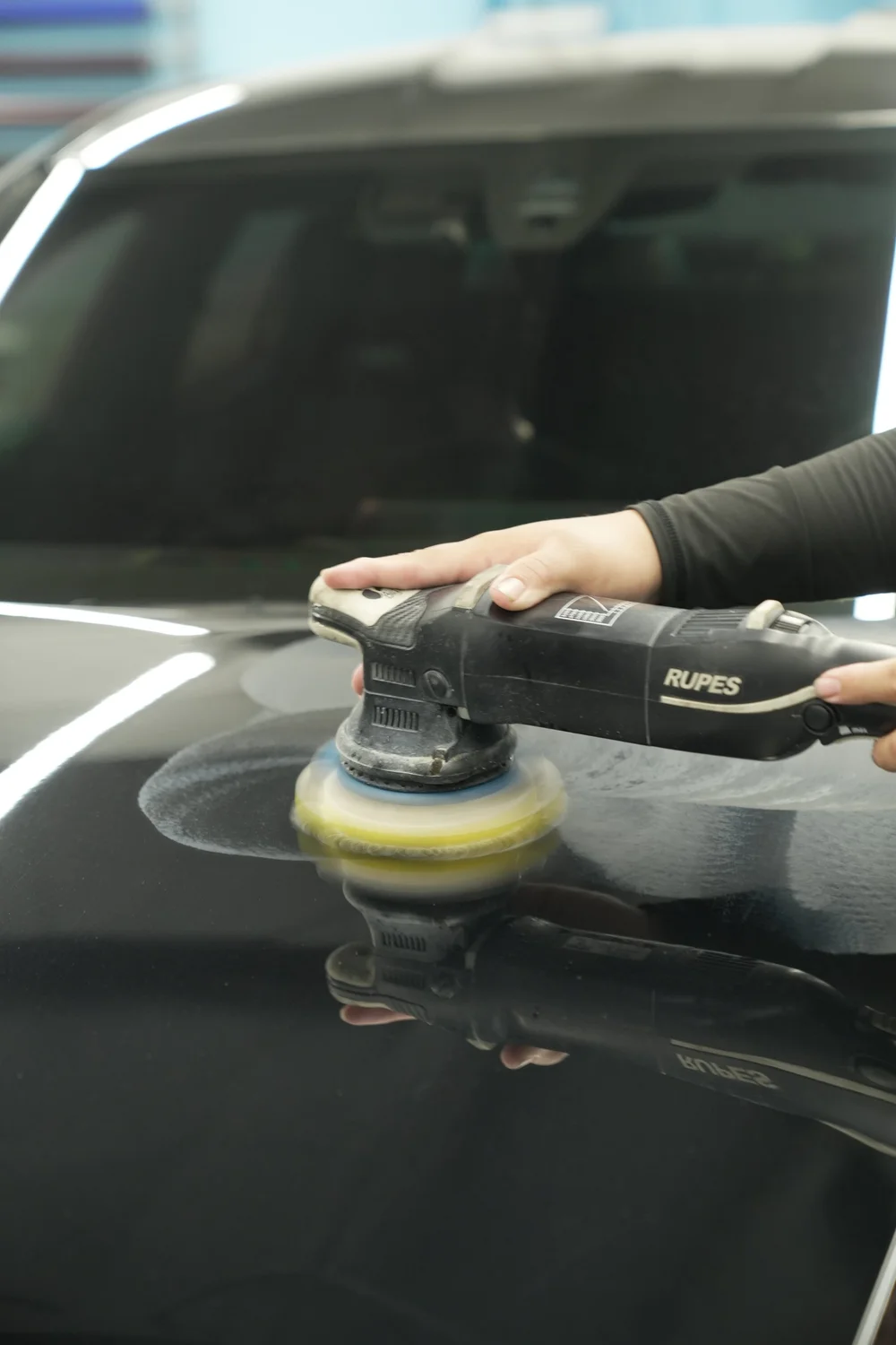 Hands operating a RUPES machine polisher with a yellow pad on reflective dark BMW M8 bodywork inside the workshop.