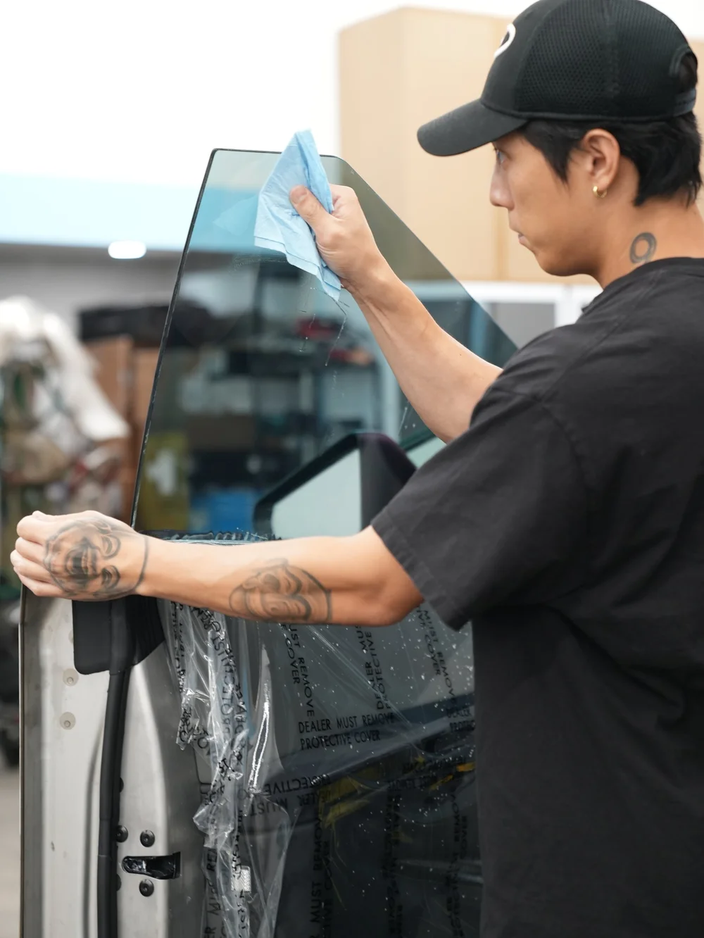 A technician wiping the upper Tesla Cybertruck side glass with a blue cloth, with an open door and protective plastic below.
