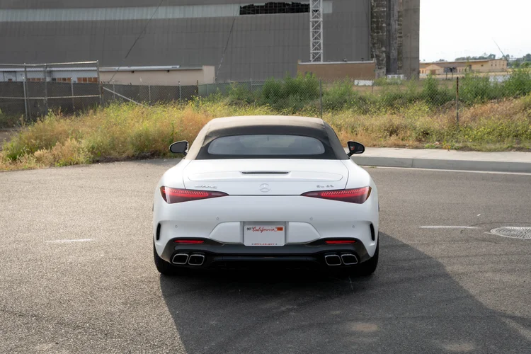 Complete higher rear view of the white SL 63 with red lamps and a black lower bumper beside grass and a gray wall