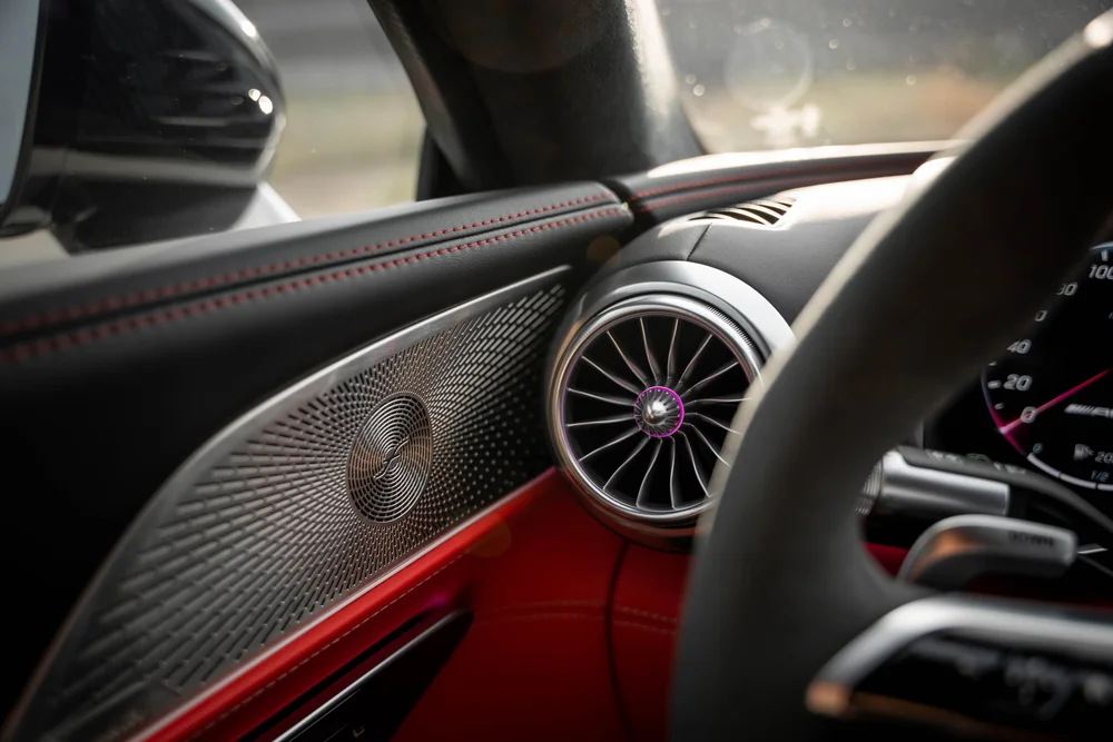 Cabin-side detail with a patterned speaker grille, circular turbine-style vent, red stitching and pink-purple light beside glass and a mirror