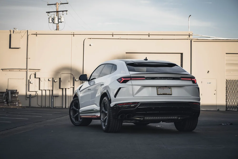 Complete rear three-quarter view of the white Urus against a beige wall, showing tricolor lower-side bands and gray wheels.