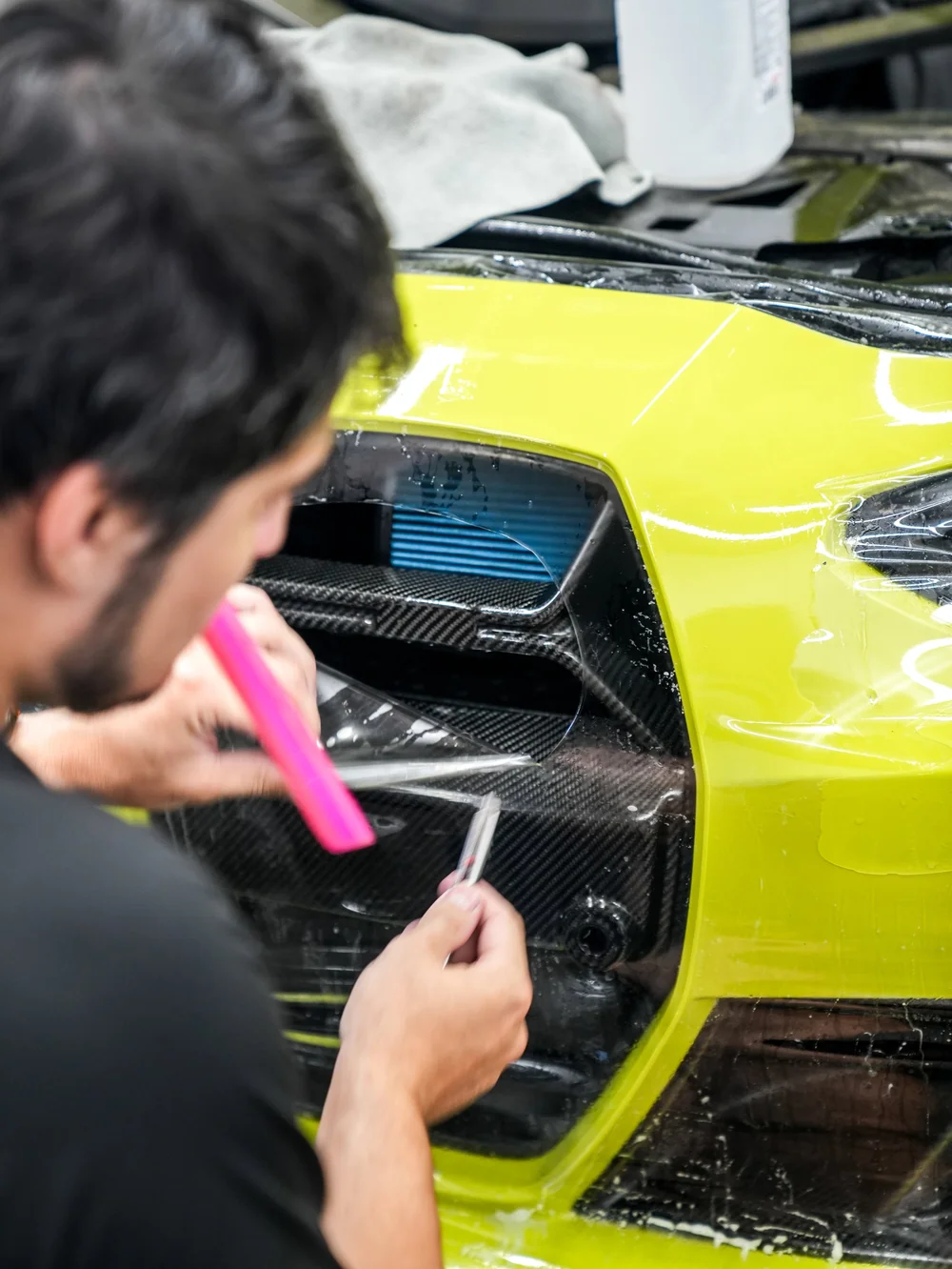 Close BMW M4 grille and front-bumper installation view showing wet transparent film, a pink flat tool and a small slender silver hand tool.