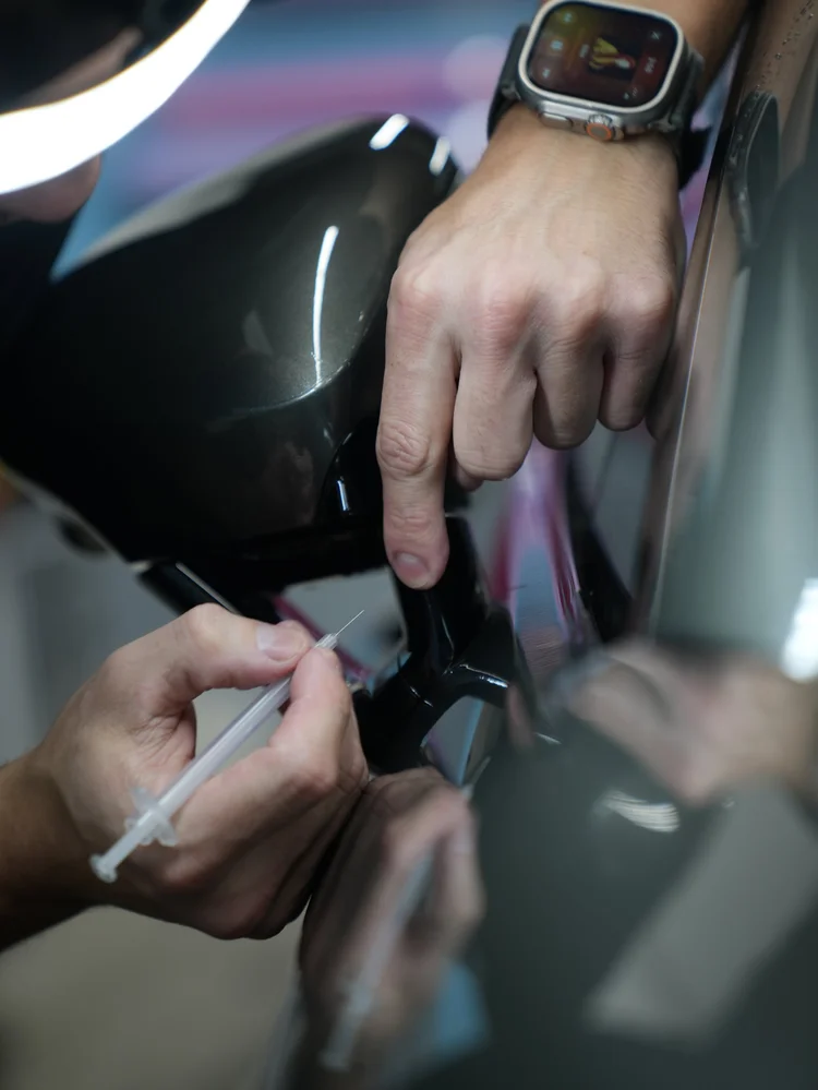 Fine needle-like applicator held at the base of a side mirror above the dark door panel
