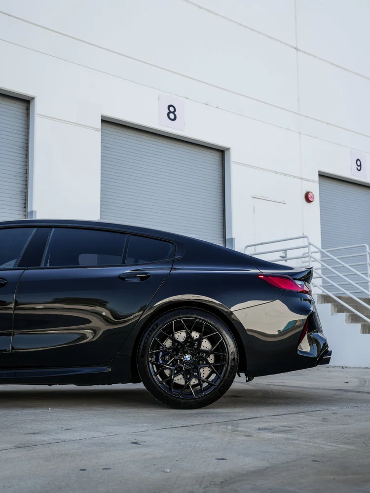 BMW M8 rear-wheel and rear-quarter detail with dark multi-spoke wheels, a blue caliper, red taillamp and reflective dark bodywork.